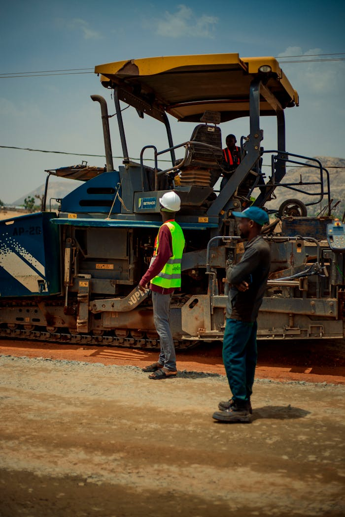 who-we-are Two construction workers observing roadwork machinery under bright daylight.